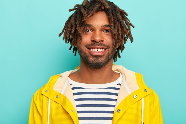 Photo gratuite happy hipster sourit agréablement, montre des dents blanches, porte un pull rayé et un imperméable jaune, heureux d'avoir une journée de congé, a marché pendant la journée d'automne, isolé sur un mur bleu