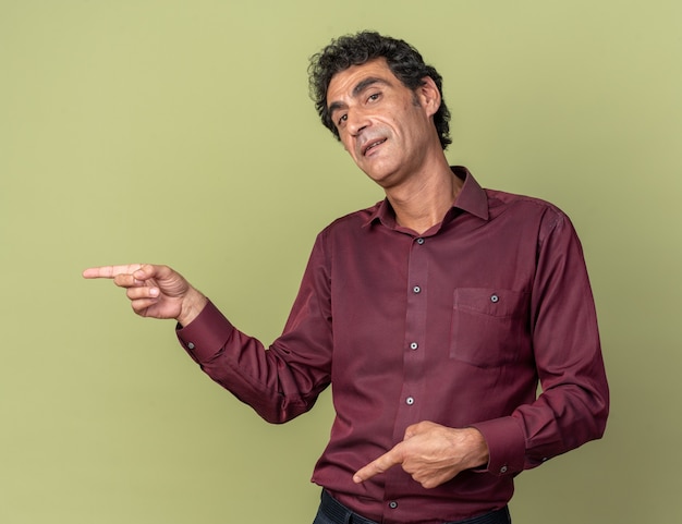 Happy senior man in purple shirt looking at camera smiling pointant avec l'index sur le côté debout sur green