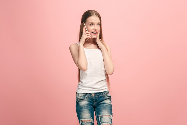 Happy teen girl debout, souriant avec un téléphone mobile sur fond de studio rose à la mode. Beau portrait de femme demi-longueur. Émotions humaines, concept d'expression faciale.