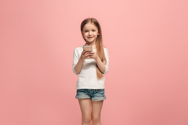 Happy teen girl debout, souriant avec un téléphone mobile sur fond de studio rose à la mode. Beau portrait de femme demi-longueur. Émotions humaines, concept d'expression faciale.