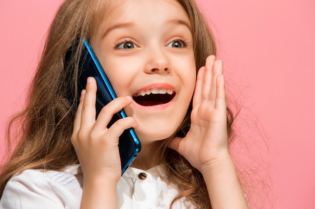 Happy teen girl debout, souriant avec un téléphone mobile sur un mur de studio rose à la mode