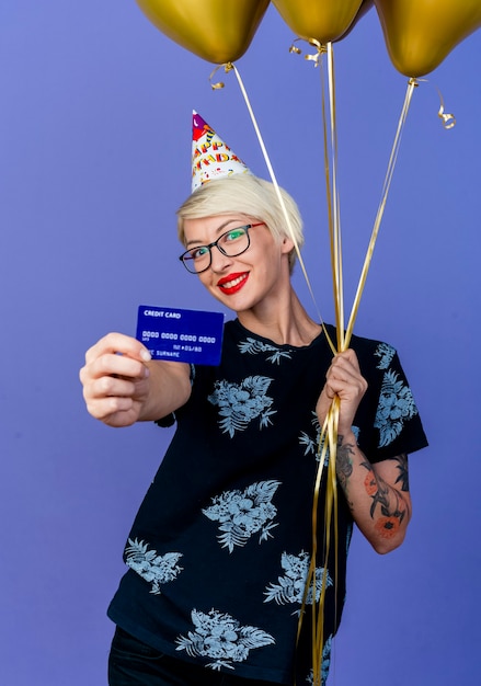 Happy young blonde party girl portant des lunettes et une casquette d'anniversaire tenant des ballons et étirant la carte de crédit regardant la caméra isolée sur fond violet