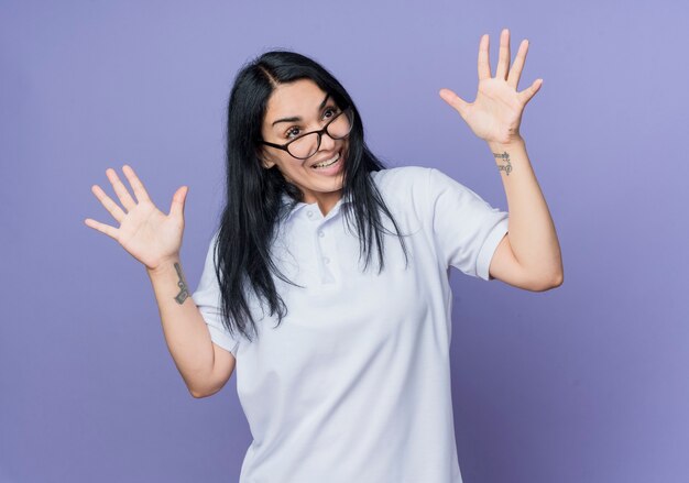 Happy young brunette caucasian girl à lunettes optiques lève les mains et regarde côté isolé sur mur violet