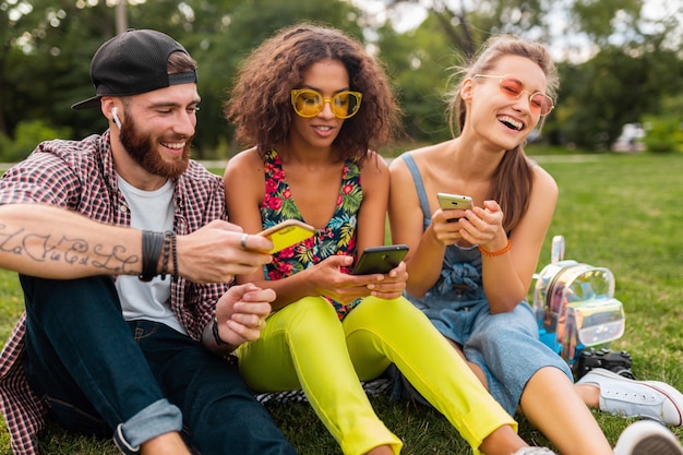 Happy young company of smiling friends sitting park à l'aide de smartphones, homme et femme s'amusant ensemble