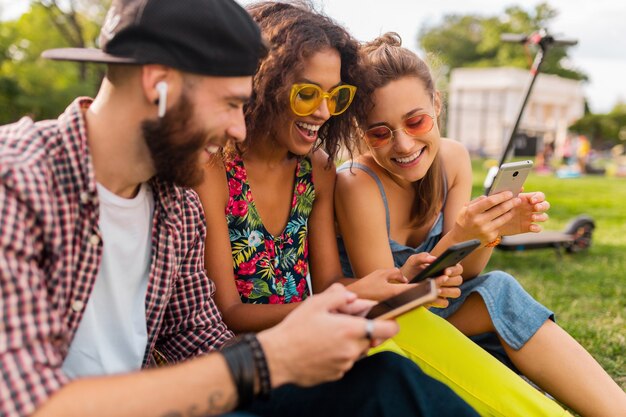 Happy young company of smiling friends sitting park à l'aide de smartphones, homme et femme s'amusant ensemble