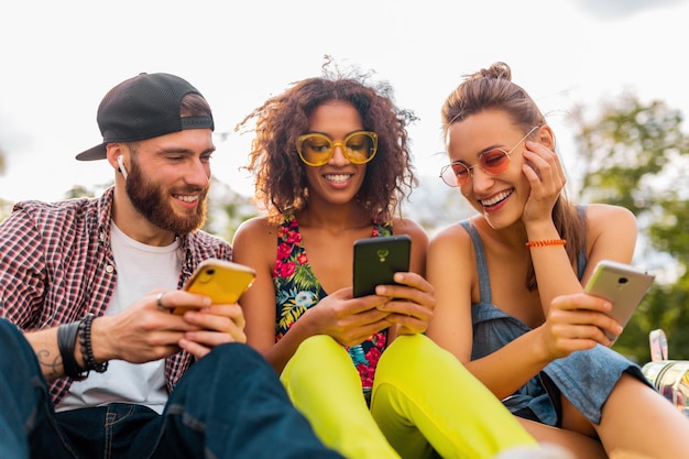 Happy young company of smiling friends sitting park à l'aide de smartphones, homme et femme s'amusant ensemble