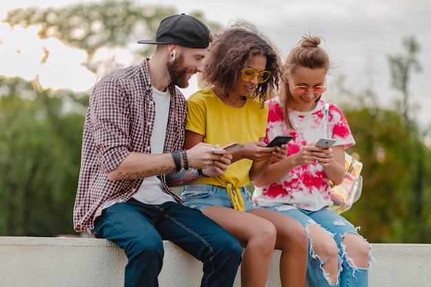 Happy young company of smiling friends sitting park à l'aide de smartphones, homme et femme s'amusant