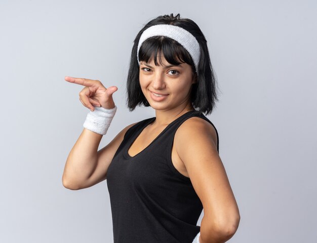 Happy young fitness girl with headband looking at camera smiling confiant pointant avec l'index sur le côté debout sur blanc