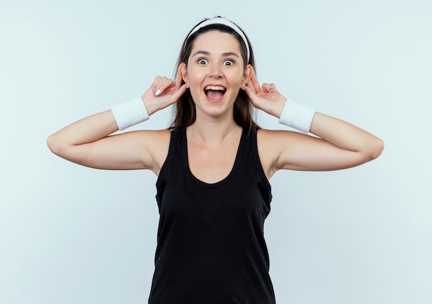 Happy young woman in headband montrant ses oreilles souriant joyeusement debout sur un mur blanc