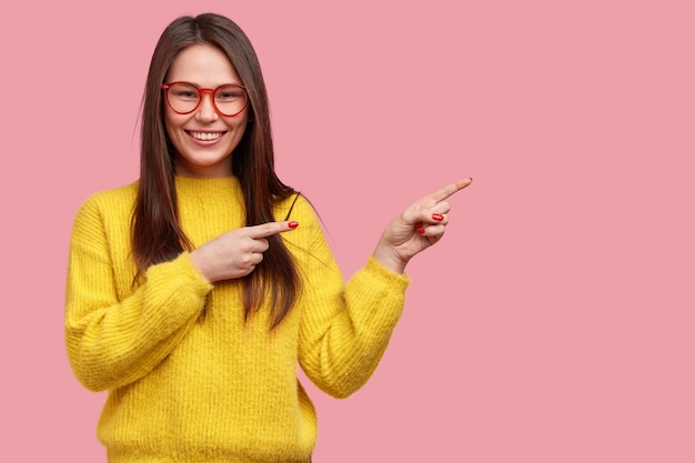 Happy young woman with straight dark hair points de côté, annonce un nouvel article dans un magasin de vêtements, habillé en pull jaune vif