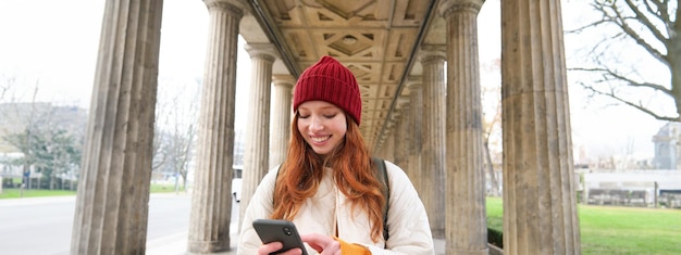 Photo gratuite le haut débit mobile et les gens souriants, une fille rousse avec un sac à dos, utilise un smartphone dans la rue