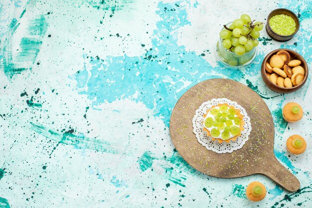 Haut de la page vue éloignée du petit gâteau avec de la crème délicieuse et des biscuits de raisins verts tranchés et frais isolés sur un bureau de lumière bleue