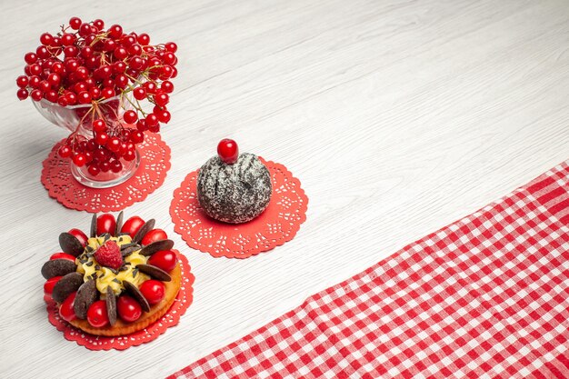 Haut de la page vue rapprochée de groseille rouge dans un gâteau aux baies en verre de cristal et gâteau au cacao sur le napperon en dentelle ovale rouge et nappe à carreaux rouge-blanc sur la table en bois blanche