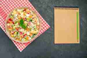 Photo gratuite haut de la page vue rapprochée de la salade de légumes dans un bol sur une serviette rouge avec bloc-notes sur le côté sur fond gris foncé