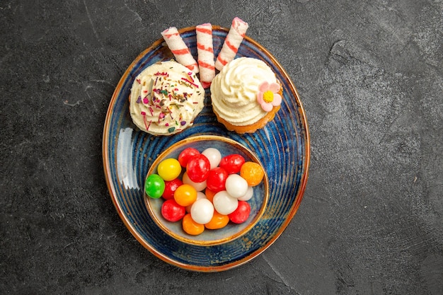 Haut vue en gros plan des bonbons sur l'assiette les appétissants cupcakes à côté du bol de bonbons colorés sur la table sombre