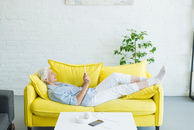 Haute femme allongée sur un canapé jaune en regardant une tablette numérique