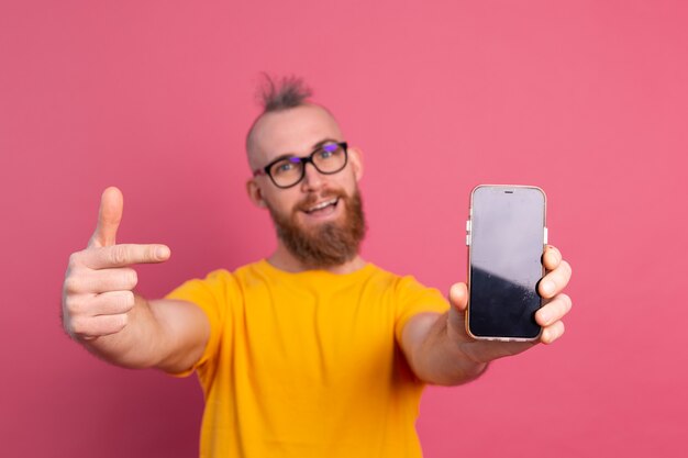 Hé quelque chose de nouveau. Heureux gars barbu européen pointant son téléphone portable avec écran blanc noir sur rose