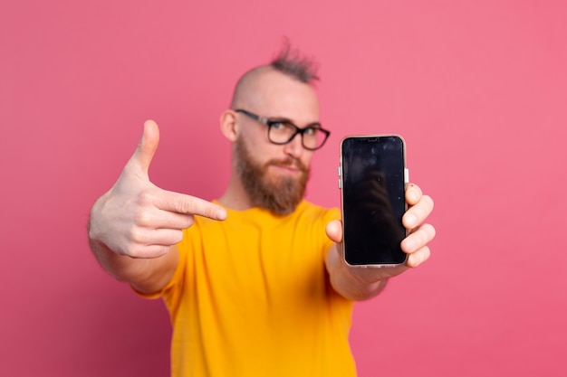 Hé quelque chose de nouveau. Heureux gars barbu européen pointant son téléphone portable avec écran blanc noir sur rose