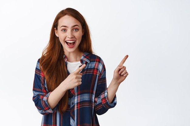 Hé, regarde ça. Femme séduisante souriante aux cheveux roux, pointant les doigts vers la droite et ayant l'air heureuse, excitée par l'offre promotionnelle, montrant la publicité, fond blanc.