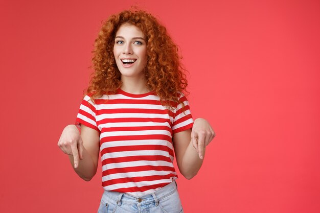 Hé, regarde ça mon ami. Sassy belle copine rousse coquine élégante coiffure frisée porte un t-shirt d'été pointant vers le bas l'index souriant amusé ravi présente une recommandation impressionnante.