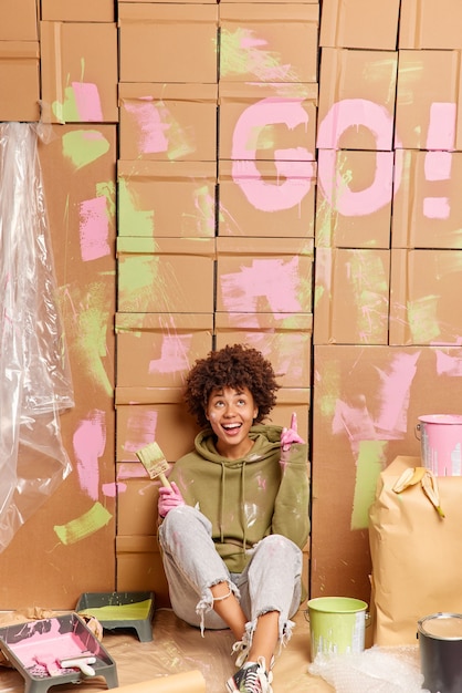 Hé regarde ce que j'ai fait! Une femme ethnique à la peau sombre et joyeuse montre comment elle a peint des murs dans un appartement entouré d'outils de peinture occupés à réparer à la maison et à redécorer la pièce