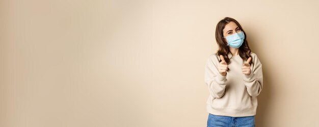 Photo gratuite hé vous félicitations belle femme souriante dans un masque médical pointant du doigt la caméra debout ov