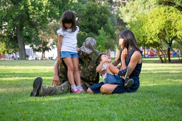 Héhé, Assis Sur L'herbe Dans Le Parc De La Ville. Père D'âge Moyen Caucasien En Uniforme Militaire, Mère Souriante Et Enfants Se Détendre Ensemble Sur La Prairie. Réunion De Famille, Week-end Et Concept De Retour à La Maison