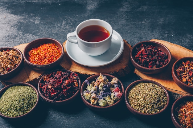 Herbes à Thé Dans Des Bols Avec Des Talons En Bois Et Une Tasse De Thé Vue En Plongée Sur Un Fond Texturé Sombre. Espace Pour Le Texte