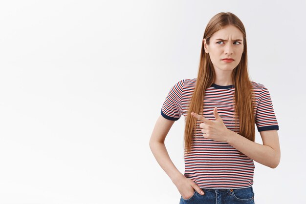 Hésitante et méfiante incertaine jeune femme blonde en t-shirt rayé fronçant les sourcils louchant incertain pointant vers la gauche et coup d'oeil sceptique debout confus fond blanc voir quelque chose d'étrange