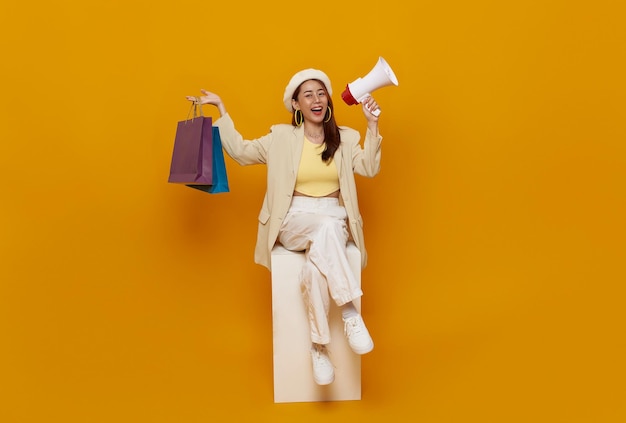 Photo gratuite heureuse adolescente asiatique assise sur une chaise tenant des sacs de shopping et annonce de réduction dans un mégaphone