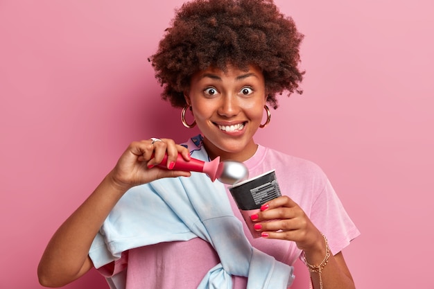 Heureuse adolescente aux cheveux afro, mord les lèvres et mange de la glace à la fraise appétissante, regarde positivement, bénéficie d'un dessert d'été froid, habillé avec désinvolture, isolé sur un mur rose