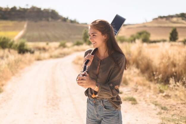 Heureuse adolescente debout sur la route de terre tenant la guitare