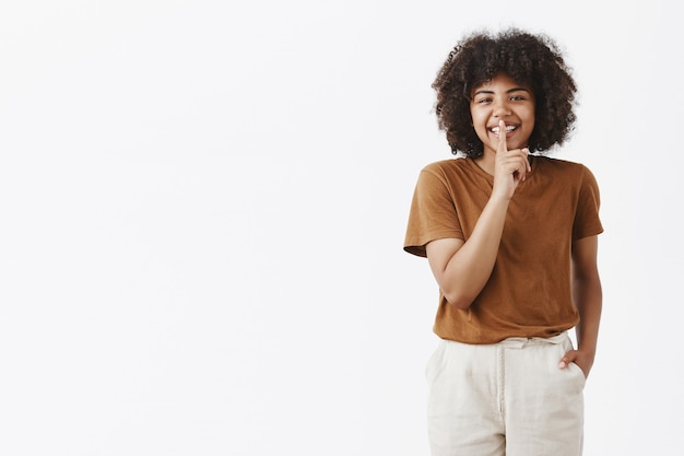 Heureuse adolescente mignonne afro-américaine insouciante avec une coiffure afro dans un t-shirt marron élégant et un pantalon blanc tenant la main dans la poche souriant avec désinvolture, montrant le signe chut avec l'index sur la bouche