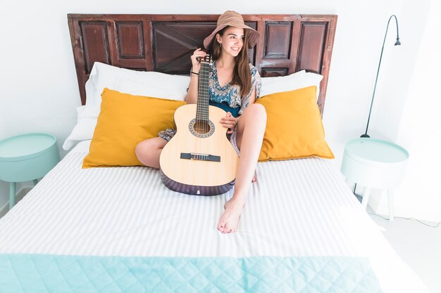 Heureuse adolescente portant chapeau assis sur le lit avec guitare