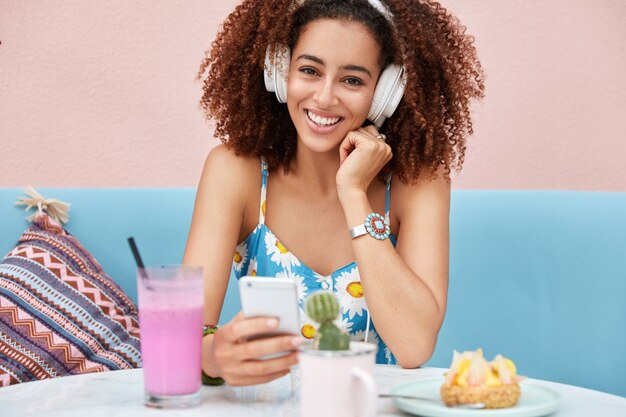 Heureuse Adorable Femme à La Peau Sombre écoute De La Musique Dans Des écouteurs, Tient Un Téléphone Intelligent, S'assoit Sur Un Canapé Confortable Dans Un Café, Boit Un Cocktail Frais.