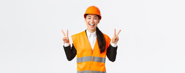 Heureuse architecte asiatique féminine optimiste en casque de sécurité et veste réfléchissante montrant le signe de la paix et souriante assurée dans la victoire de son entreprise remportant l'appel d'offres sur les travaux de construction