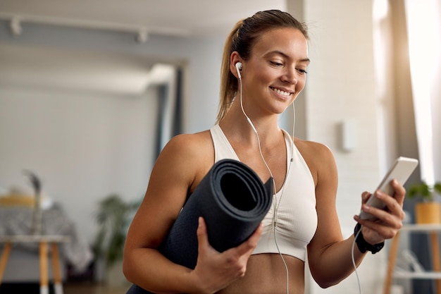 Heureuse athlète féminine tenant un tapis d'exercice tout en envoyant des SMS sur son téléphone portable à la maison