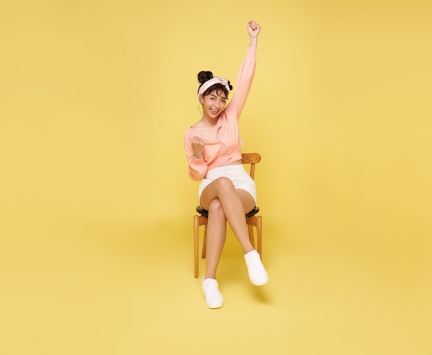 Heureuse belle adolescente asiatique assise sur une chaise et les mains en l'air célébrant le geste sur le mur jaune. Concept de réussite du gagnant excité.