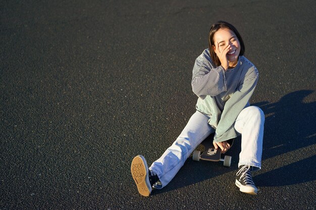 Heureuse belle adolescente coréenne est assise sur sa planche à roulettes en croisière sur longboard portant des vêtements décontractés