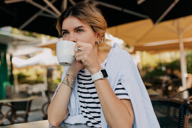 Heureuse belle femme aux cheveux clairs portant un t-shirt rayé et une chemise bleue est assise sur un café d'été et boit du café le matin au soleil