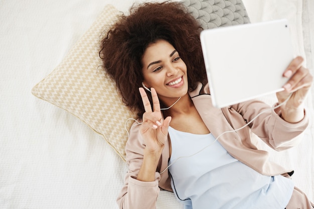 Photo gratuite heureuse belle femme dans les écouteurs souriant montrant la paix faisant selfie sur tablette couché dans son lit. d'en haut.