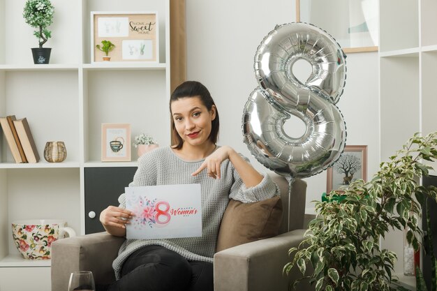 Heureuse belle femme le jour de la femme heureuse tenant et points à la carte postale assise sur un fauteuil dans le salon