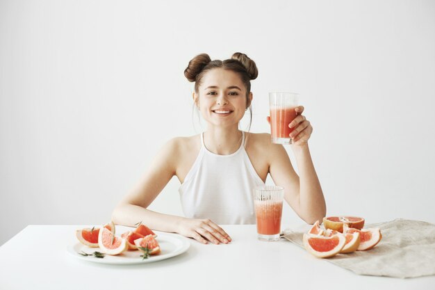 Heureuse belle femme souriante assise à table tenant le verre avec smoothie de pamplemousse frais de désintoxication saine sur le mur blanc.