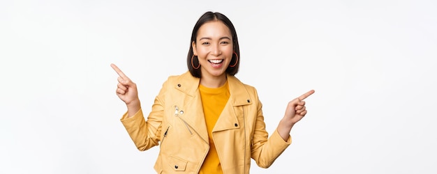 Heureuse belle fille asiatique pointant les doigts à gauche et à droite montrant le logo de la bannière démontrant la vente debout dans une veste jaune sur fond blanc