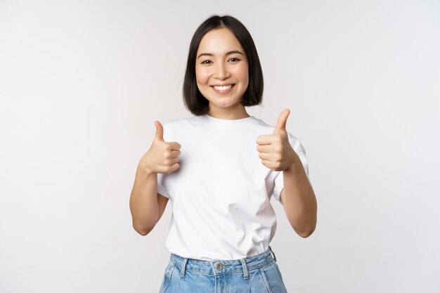 Heureuse belle fille coréenne montrant les pouces vers le haut en signe d'approbation souriant heureux comme qch recommandant debout sur fond blanc