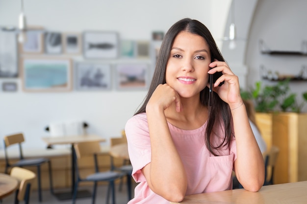 Heureuse belle jeune femme confiante parlant sur cellule, debout à co-working, se penchant sur le bureau,