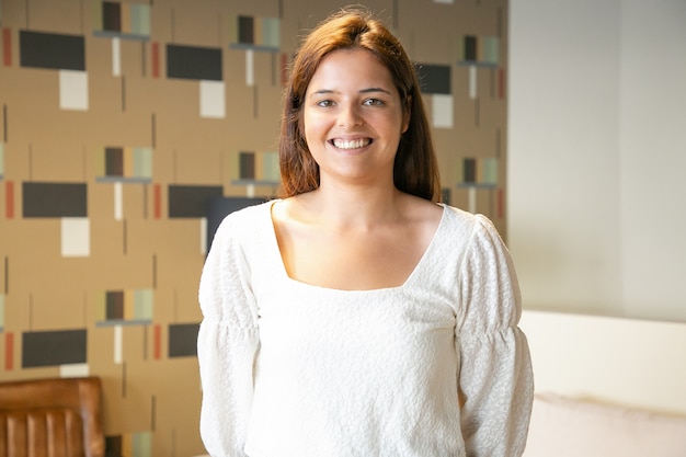 Heureuse belle jeune femme debout et posant dans l'intérieur de co-working ou de café, regardant la caméra et souriant