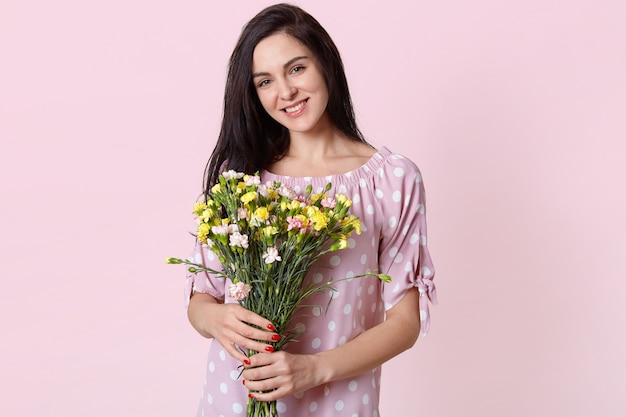 heureuse belle jeune femme européenne avec un sourire doux, porte une robe à pois, détient un bouquet de fleurs, heureuse de recevoir de son mari, des modèles sur pastel rose.
