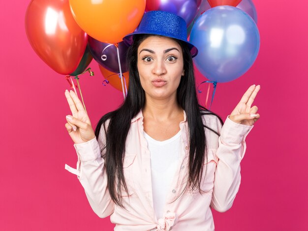 Heureuse belle jeune femme portant un chapeau de fête debout devant des ballons pointant sur différents côtés isolés sur un mur rose