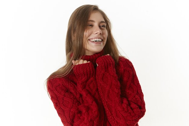 heureuse belle jeune femme portant un pull à manches longues marron, smilijg joyeusement. Fille sympathique positive avec un large sourire charmant, posant à l'intérieur dans un pull en tricot élégant